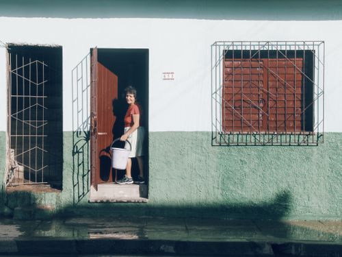 Free A woman stands in a sunlit doorway holding a bucket on a village street. Stock Photo