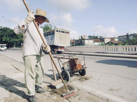 A city street cleaner in a hat sweeps the sidewalk on a sunny day with traffic in the background.