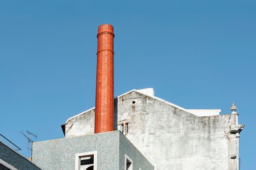 Free Low-angle view of a red-brick industrial chimney with a historical building facade against a clear blue sky. Stock Photo