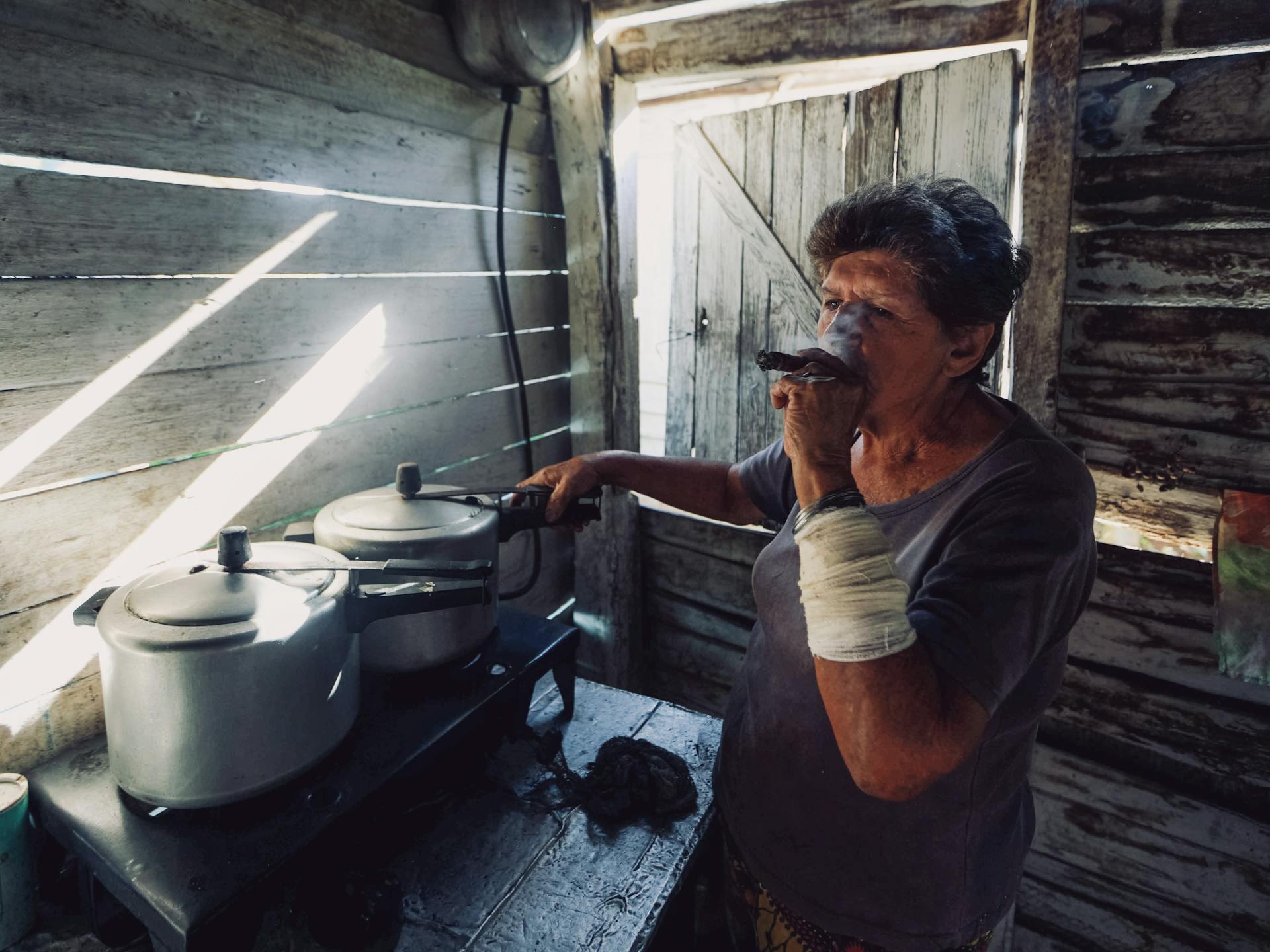 Elderly woman smoking a cigar beside metal pots on a rustic stove inside a wooden kitchen.