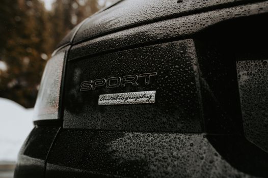 Detailed close-up of a black car with raindrops on the 'Sport' emblem, showcasing texture.