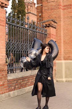 Stylish woman in black coat holding balloons near brick building outdoors.