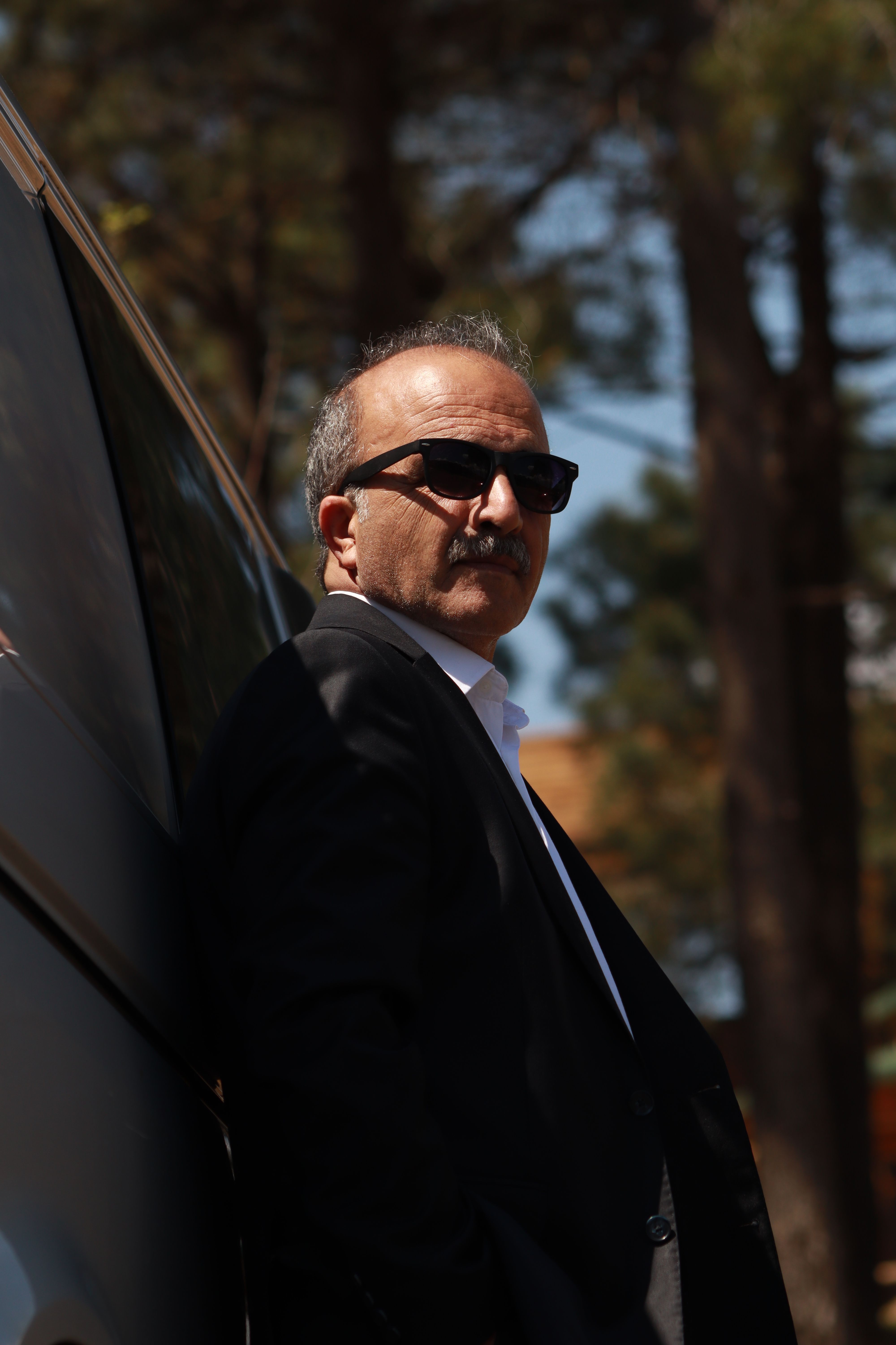 Senior man in a suit wearing sunglasses, posing confidently outdoors by a vehicle.