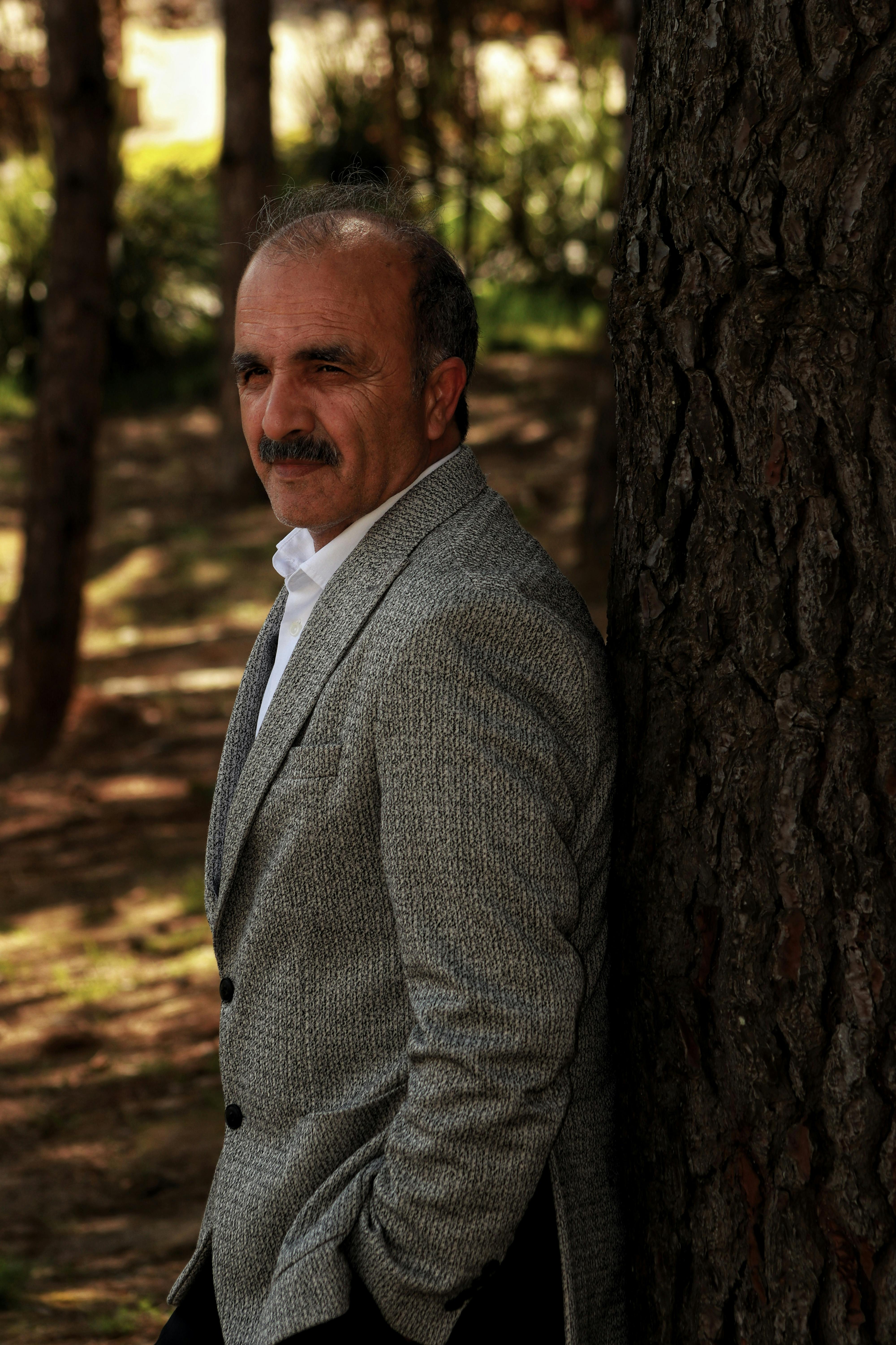 Portrait of a mature man with mustache in a grey suit, leaning against a tree outdoors.
