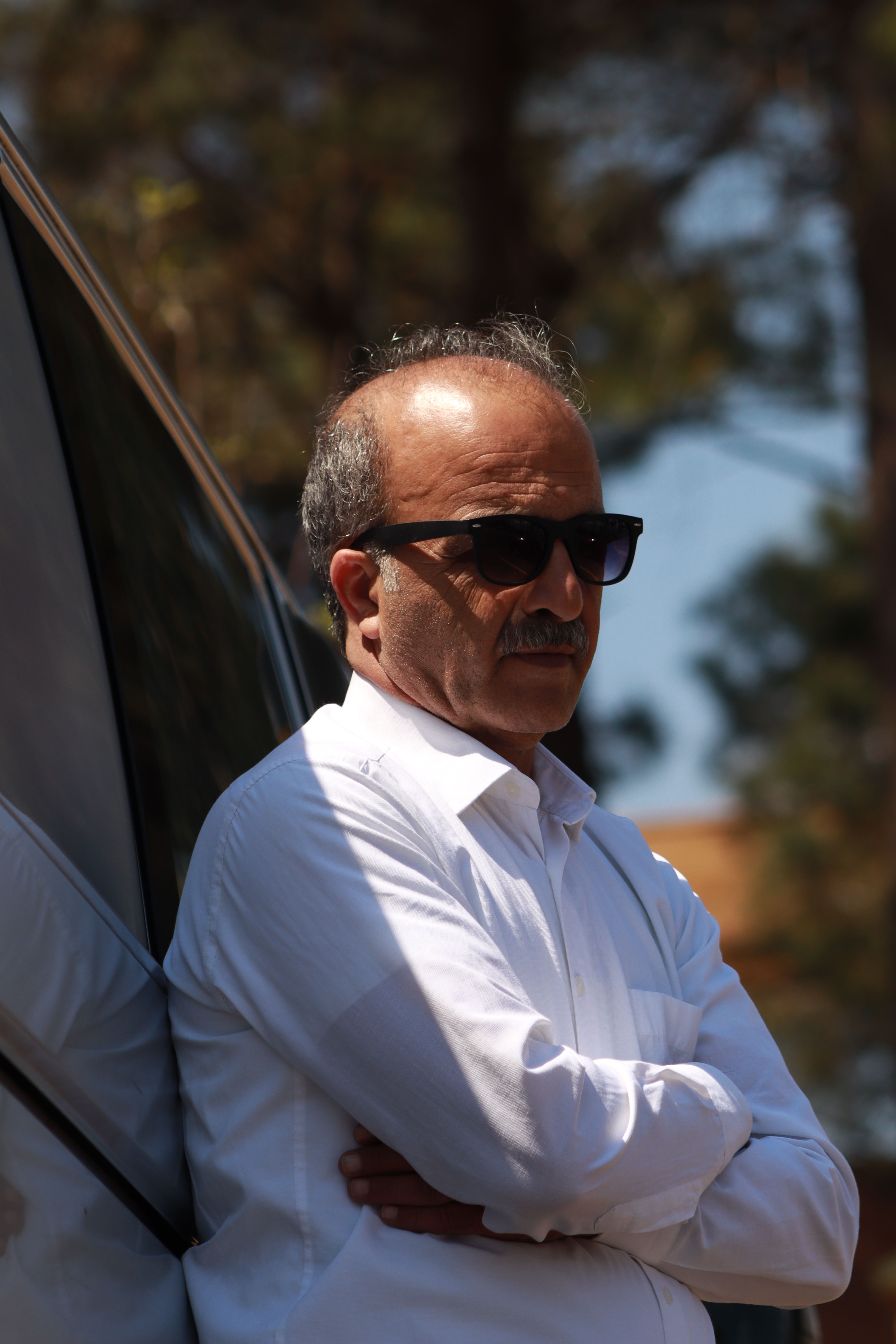 Elderly man with sunglasses poses confidently by car outdoors, wearing a white shirt.