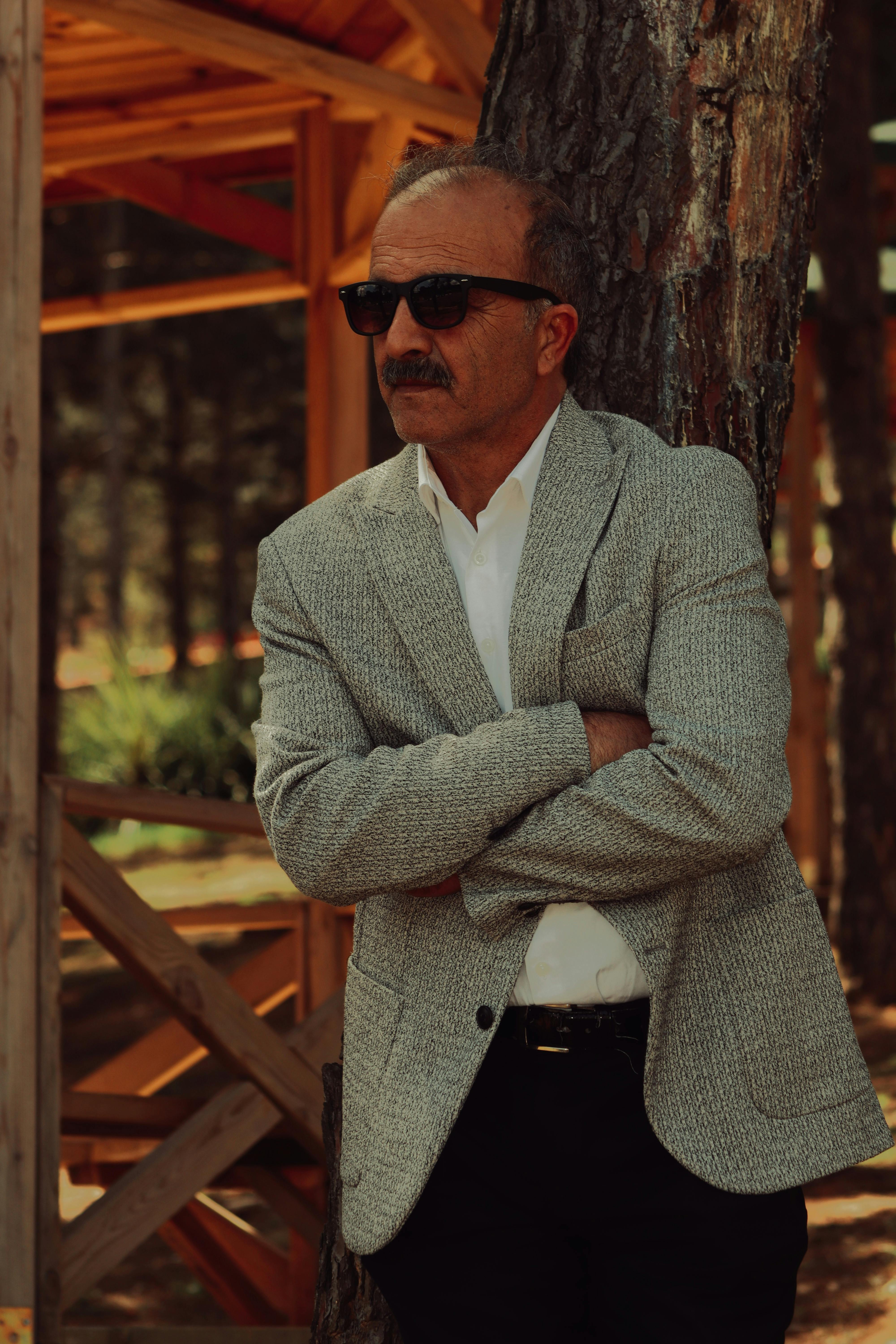 Sophisticated mature man in sunglasses posing outdoors in a grey jacket and white shirt.