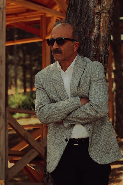 Mature Man in Grey Jacket and Sunglasses Standing by a Tree with Arms Crossed