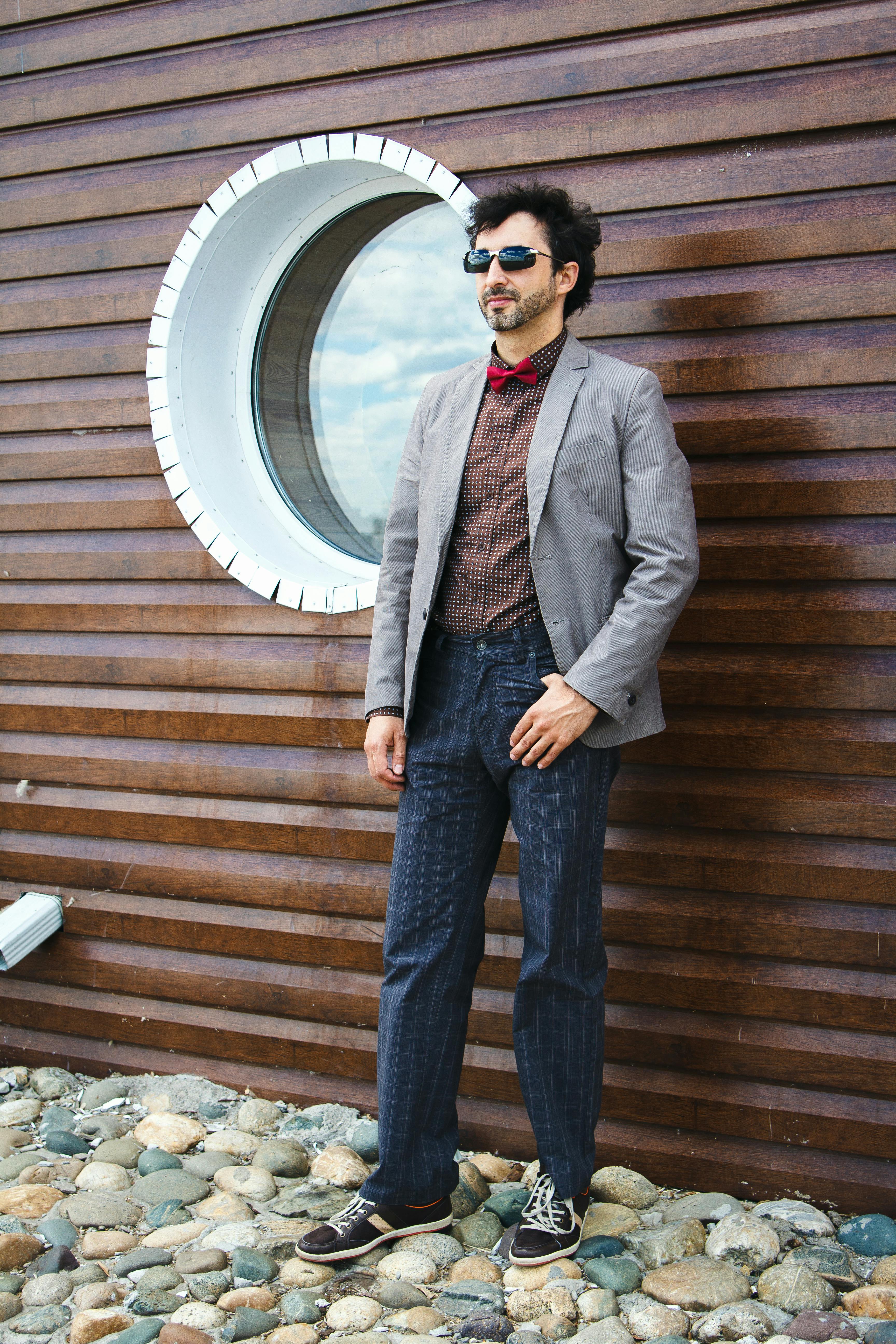 一位戴着墨镜、身穿西装外套的时尚男士，在木墙的圆形窗边摆着姿势