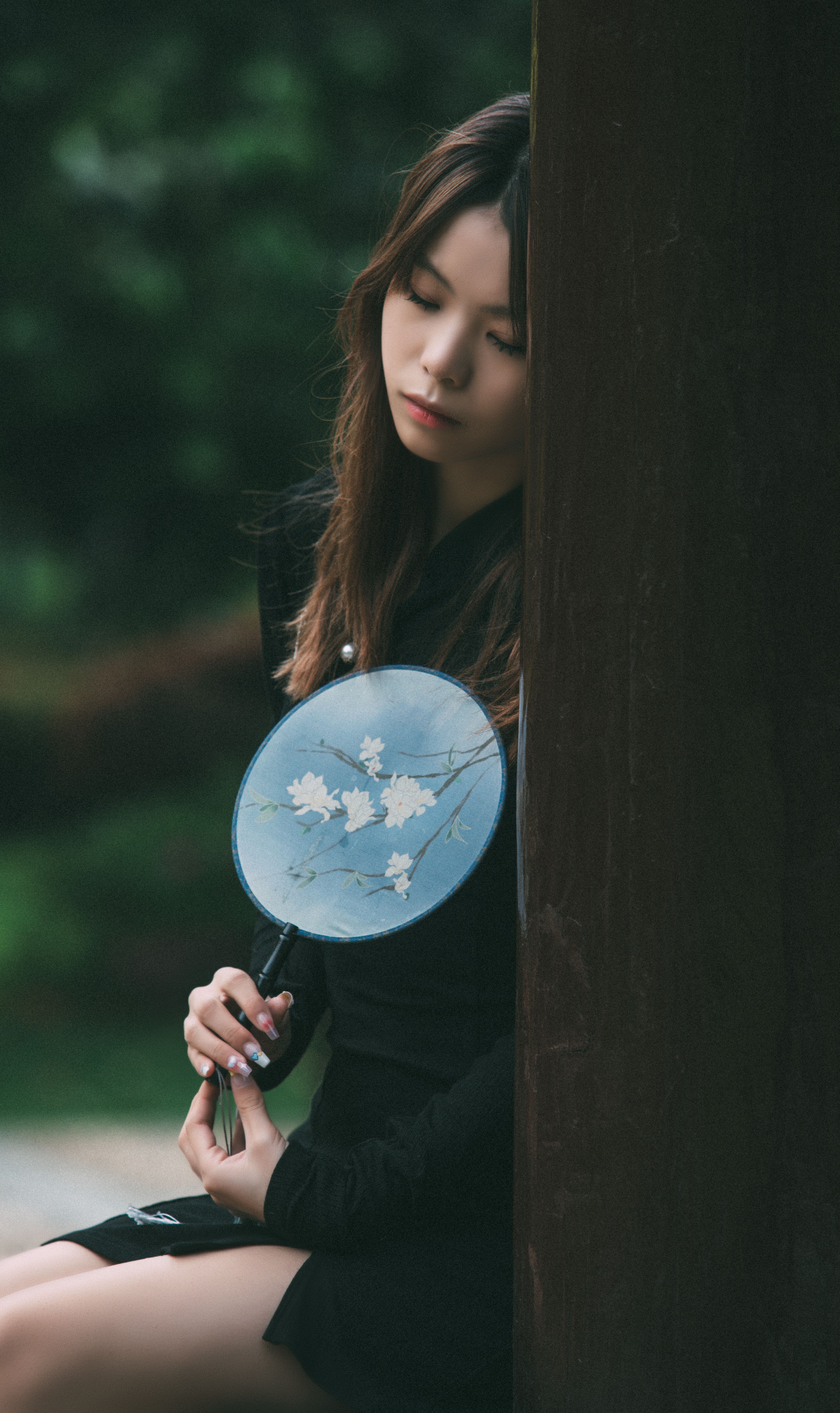 A young woman leans against a post in a serene park, holding a decorative fan, reflecting a moment of contemplation.