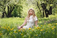 一位金发红唇的女子坐在郁郁葱葱的田野上，四周蒲公英盛开，在户外享受着夏日时光