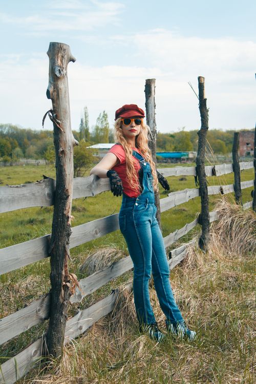 Free Blonde woman in casual overalls and cap near rustic fence on a sunny day. Stock Photo