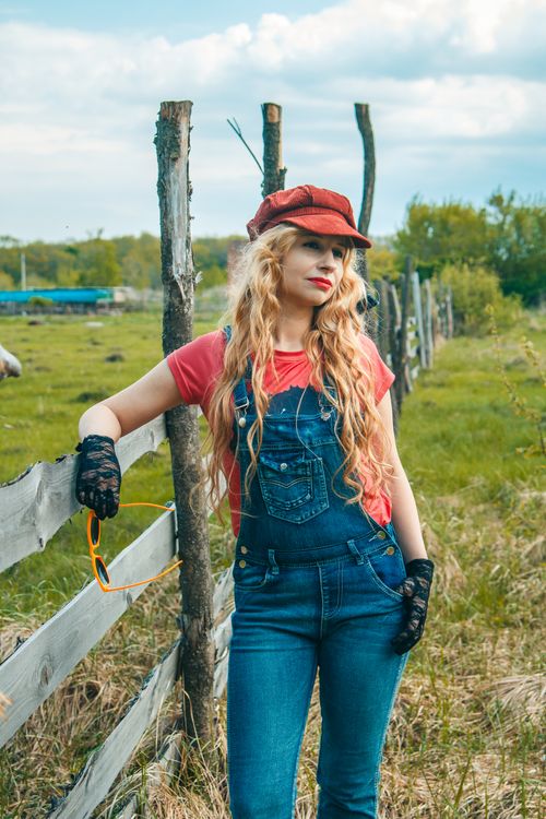 Free Fashionable young woman in casual overalls posing against a rural fence on a sunny day. Stock Photo