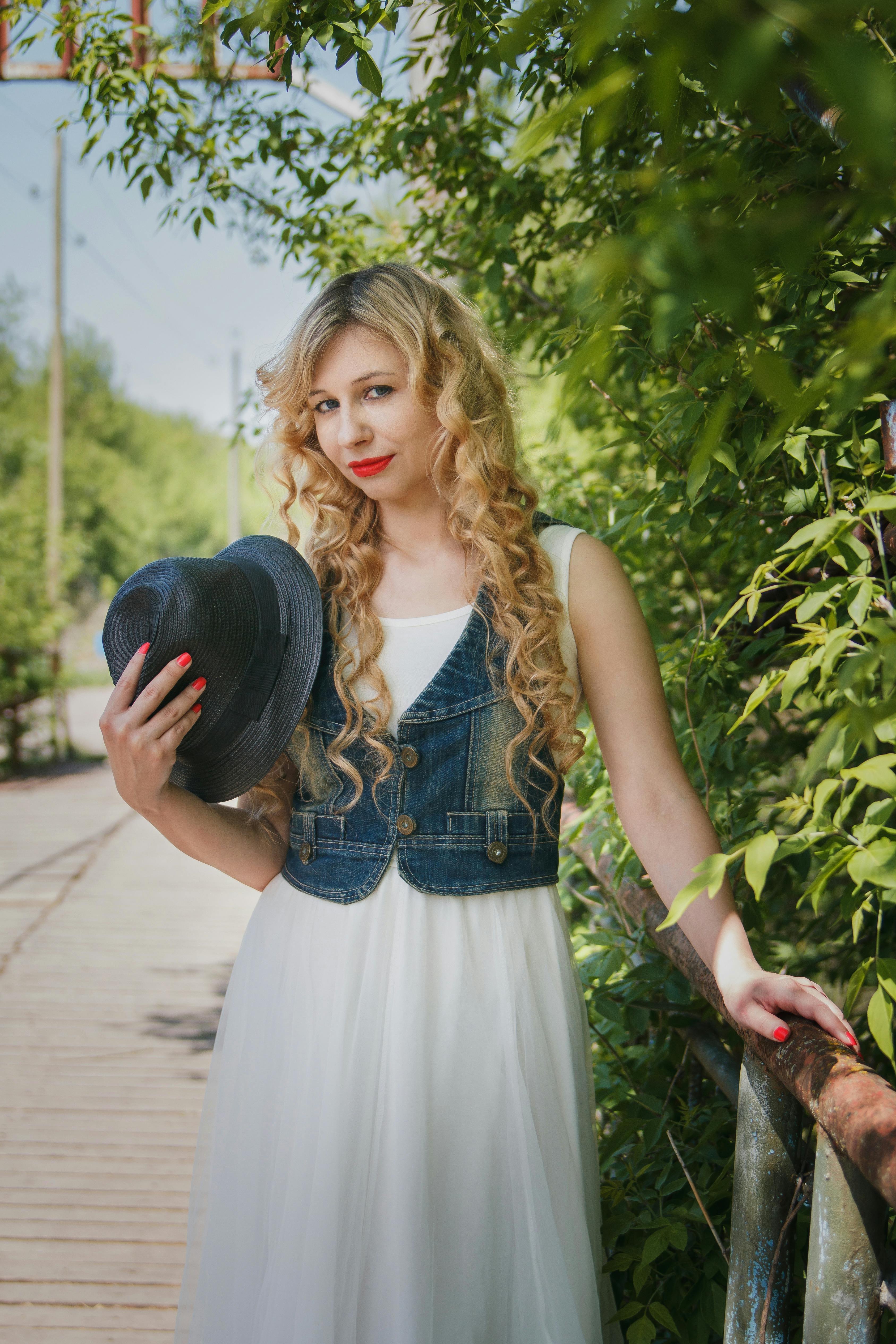 一位优雅的年轻女子在郁郁葱葱的绿色户外背景中，手拿帽子，身穿牛仔背心