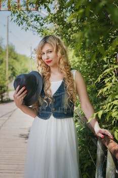 一位优雅的年轻女子在郁郁葱葱的绿色户外背景中，手拿帽子，身穿牛仔背心