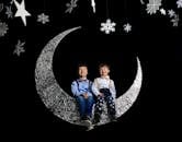 两个微笑的孩子坐在一个银色的新月上，新月装饰着雪花和星星，在一个异想天开的工作室里拍摄