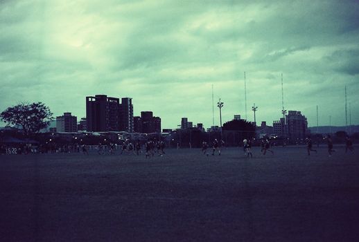 A glimpse of an urban soccer game with players running in front of city buildings under an overcast sky.