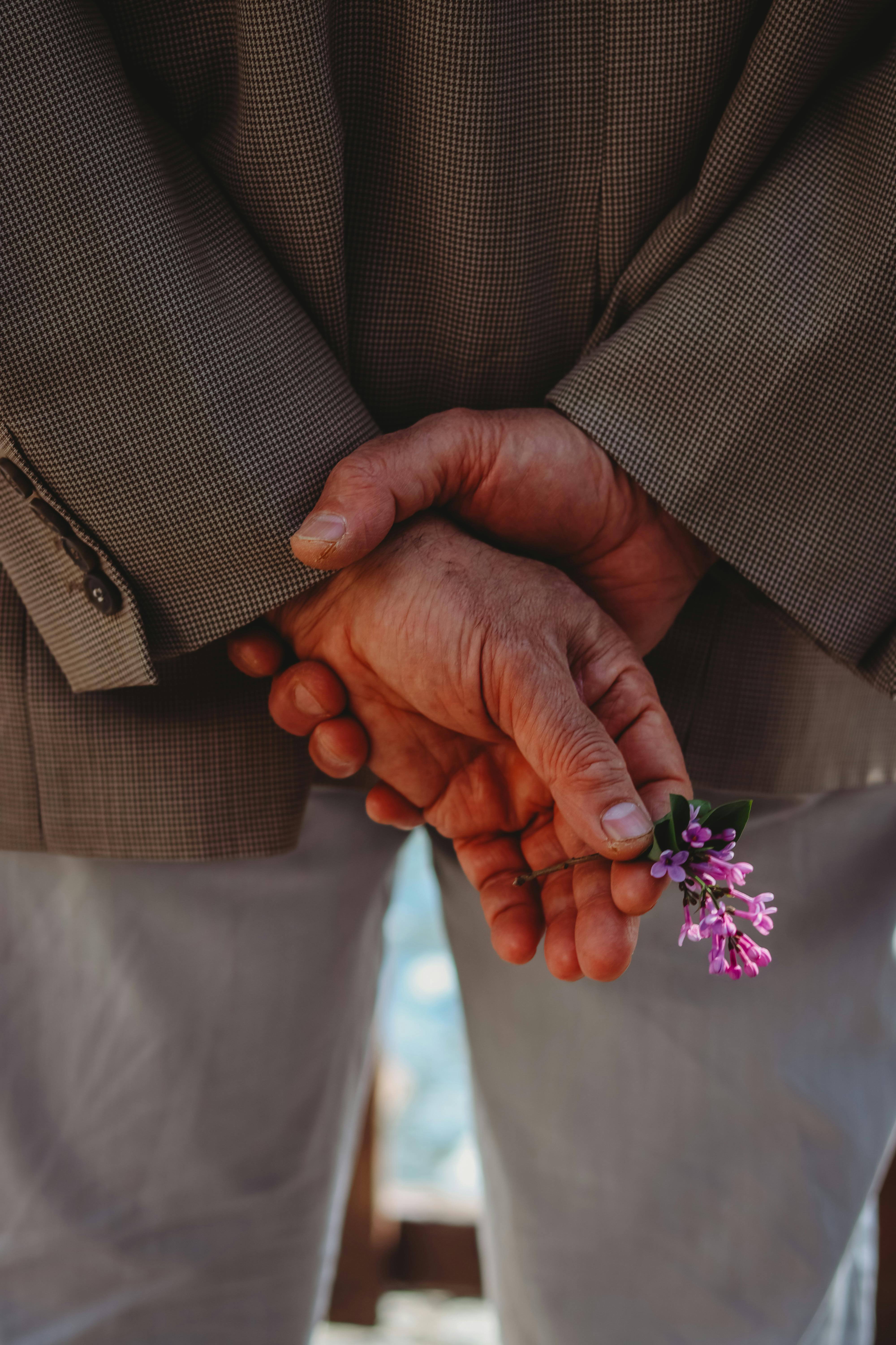A senior man in a grey jacket holds a small purple flower behind his back outdoors.