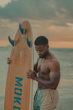 A shirtless male surfer holding a surfboard by the sea at sunset, embodying beach lifestyle.
