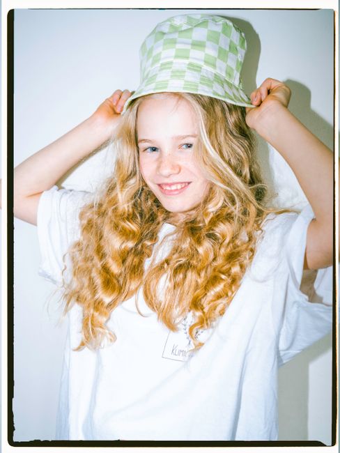 Portrait of a young girl with curly hair, wearing a green checkered hat and white t-shirt, smiling indoors.