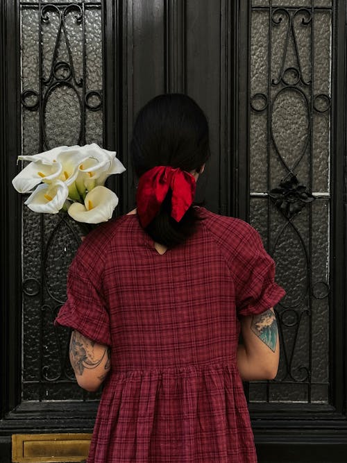 Back View of a Woman in a Checkered Dress and Holding a Bunch of Flowers Standing next to the Door 