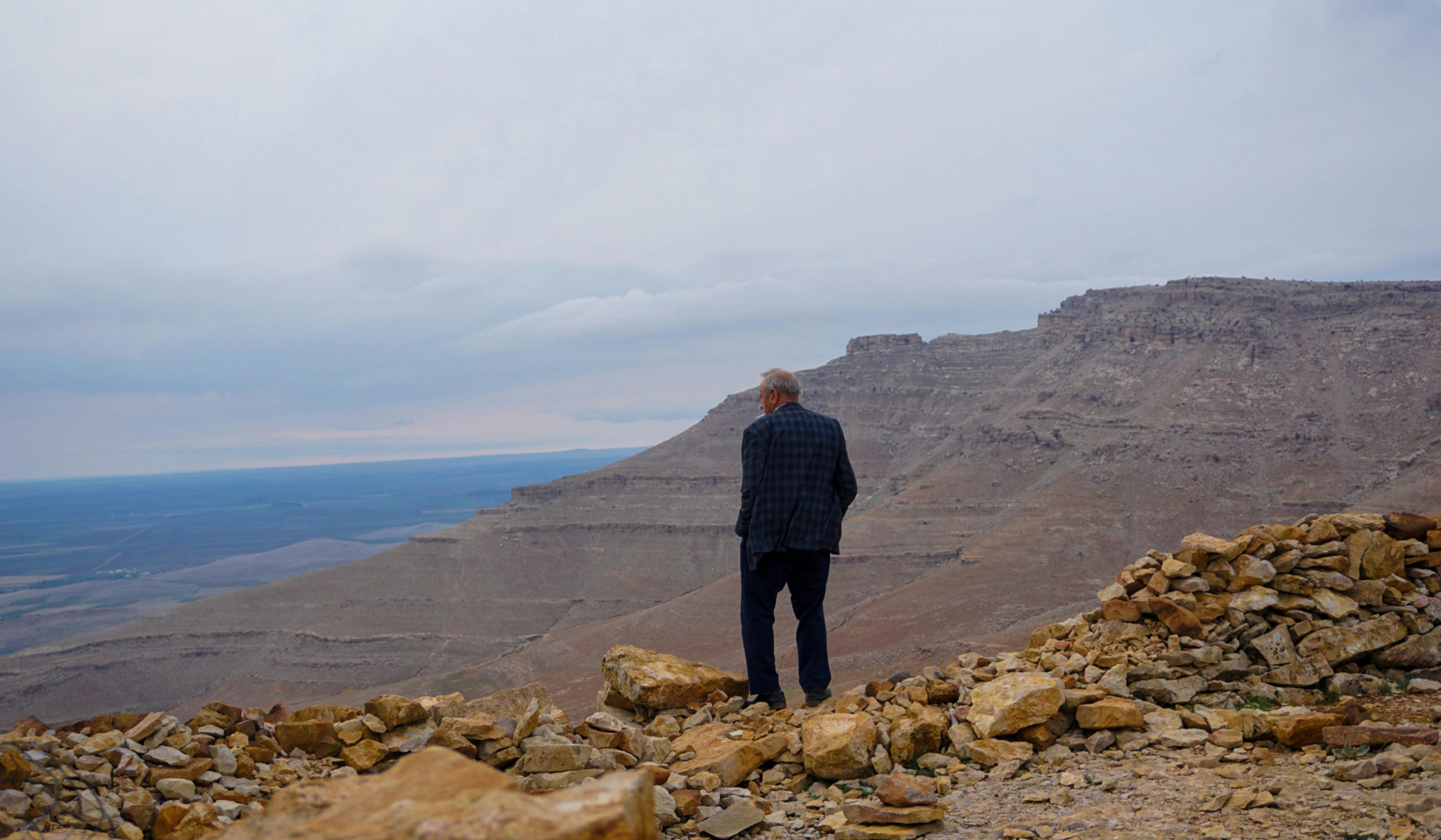 A lone man stands on rocky terrain gazing over expansive mountains and distant horizon.