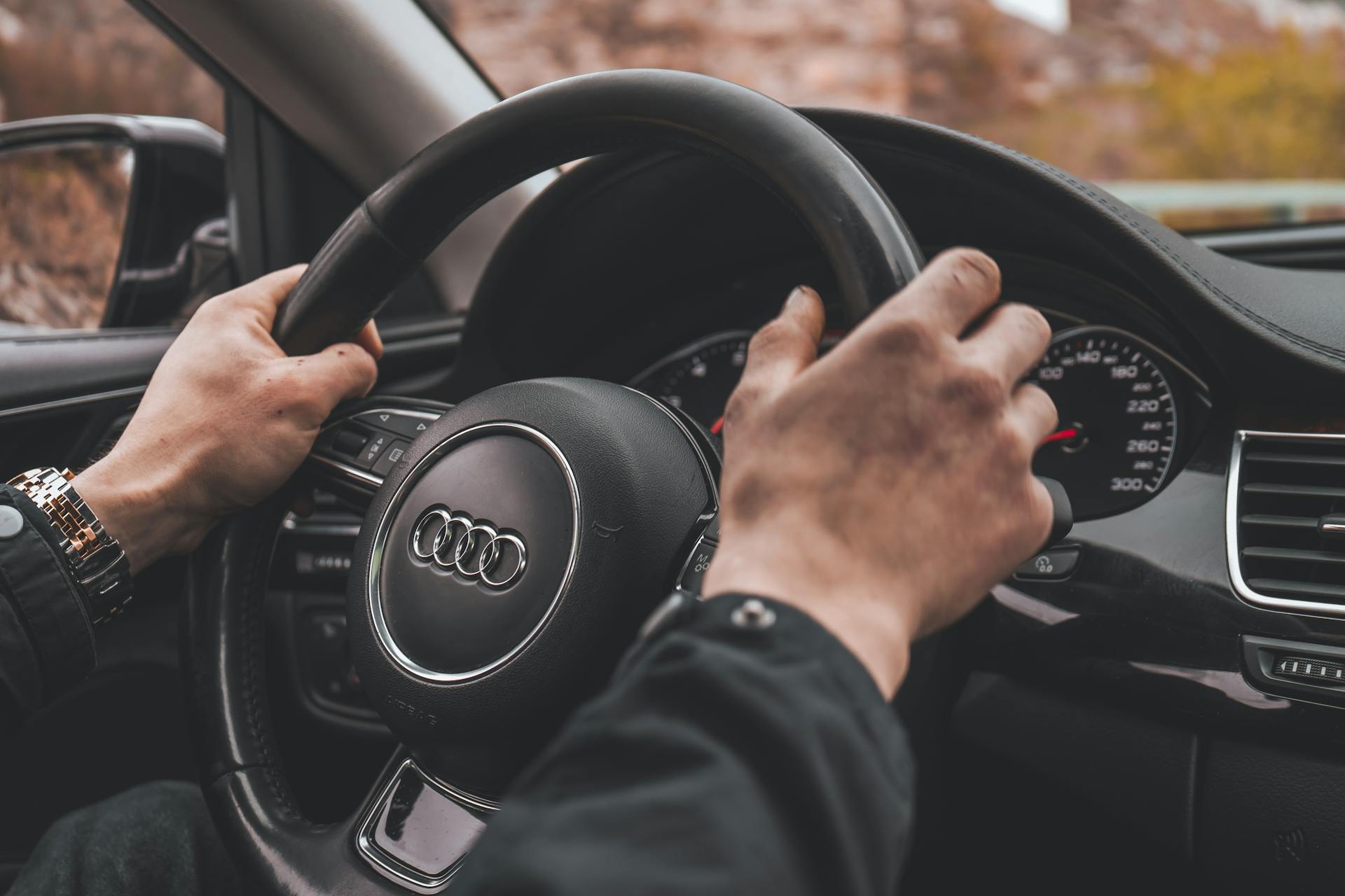 Driver's hands holding an Audi steering wheel, focusing on the car interior and gauges.