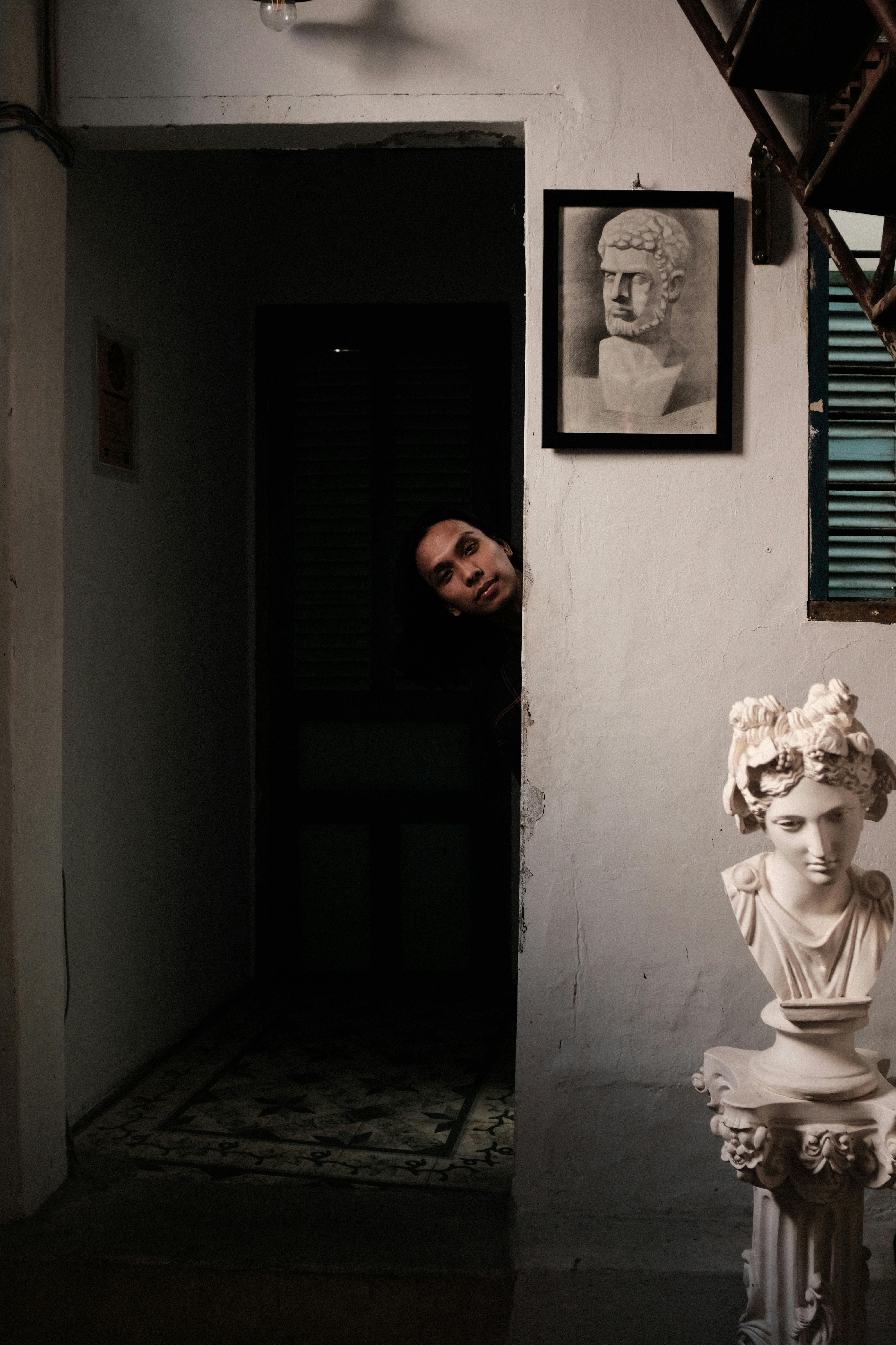 A man peeks from a dimly lit doorway next to a classical sculpture, creating a mysterious atmosphere.