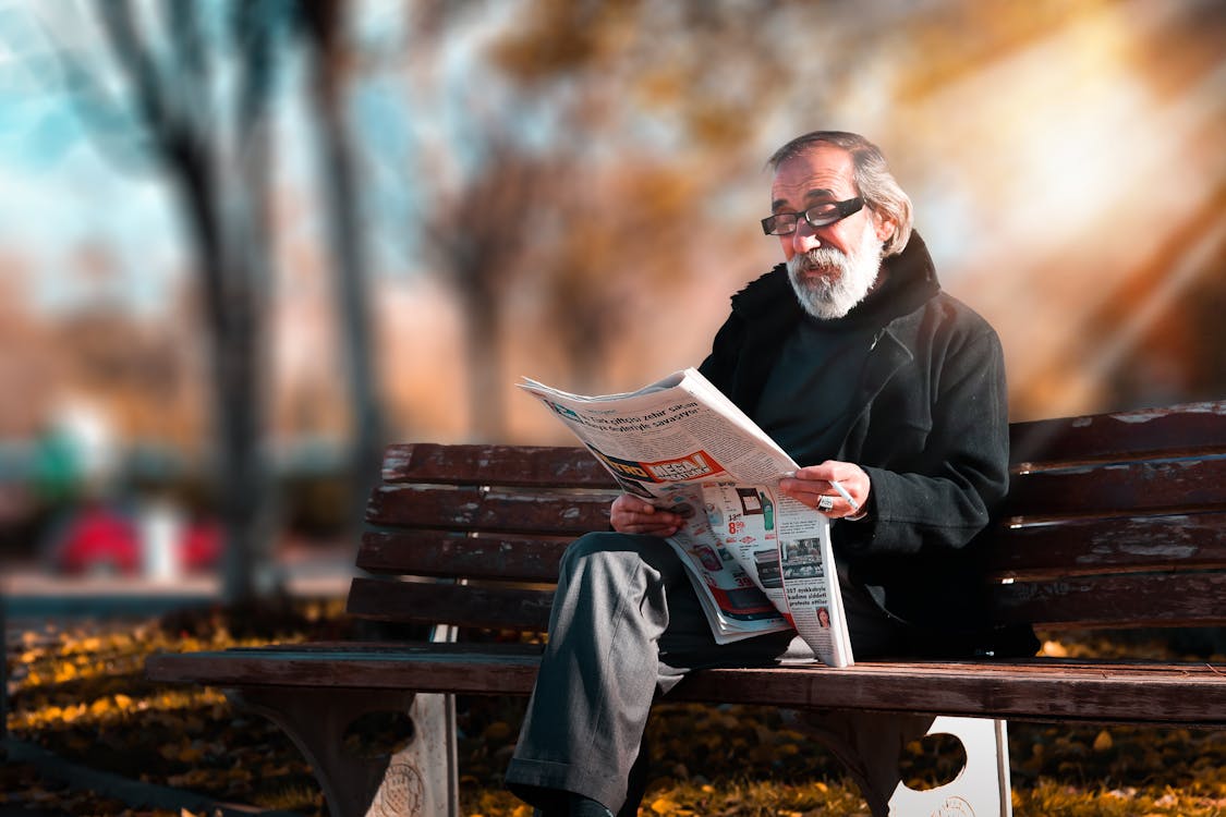 An elderly person reading a newspaper on a park bench