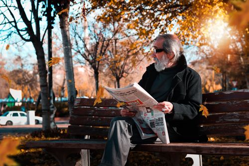 Free Senior adult enjoying a fall day outdoors while reading a newspaper on a park bench surrounded by autumn leaves. Stock Photo