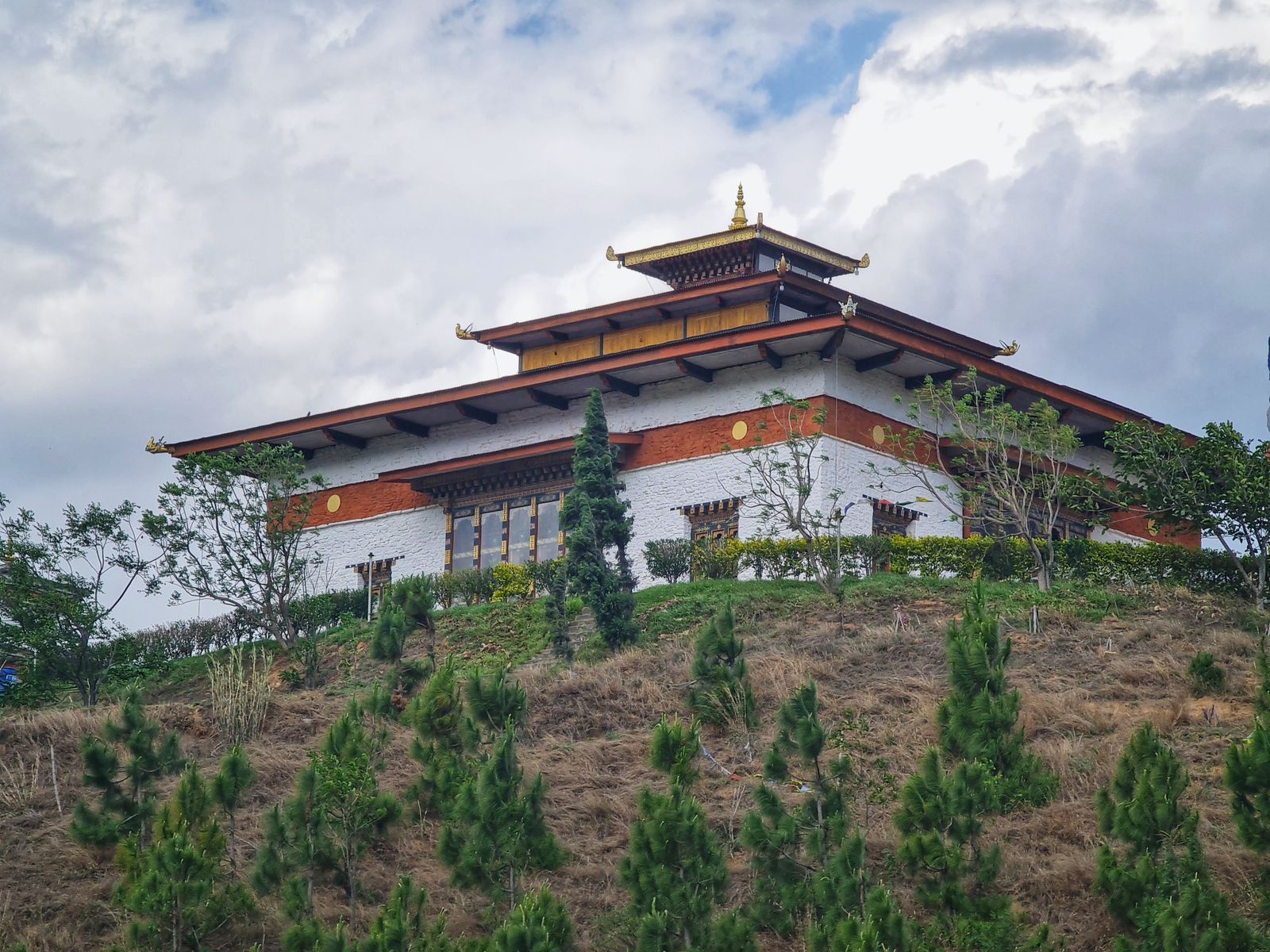 A whitewashed hilltop temple with a gilded roof among young pines in Wangdue Phodrang, cloud gathering overhead