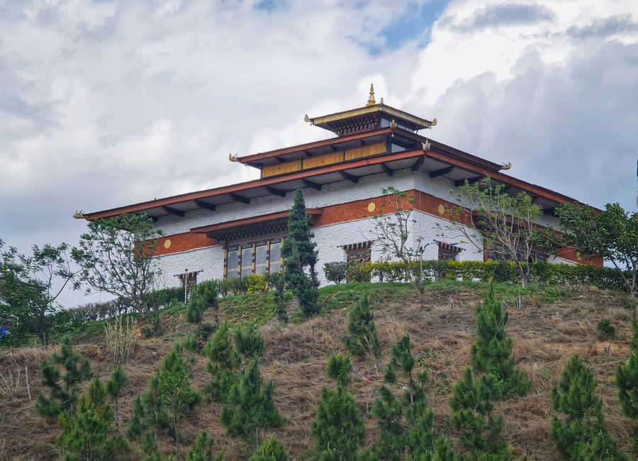 Elevated view of a traditional Bhutanese temple atop a green hillside under a cloudy sky in Wangdue Phodrang.