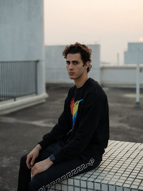 Youthful man in a casual black outfit seated on an urban rooftop during the day.