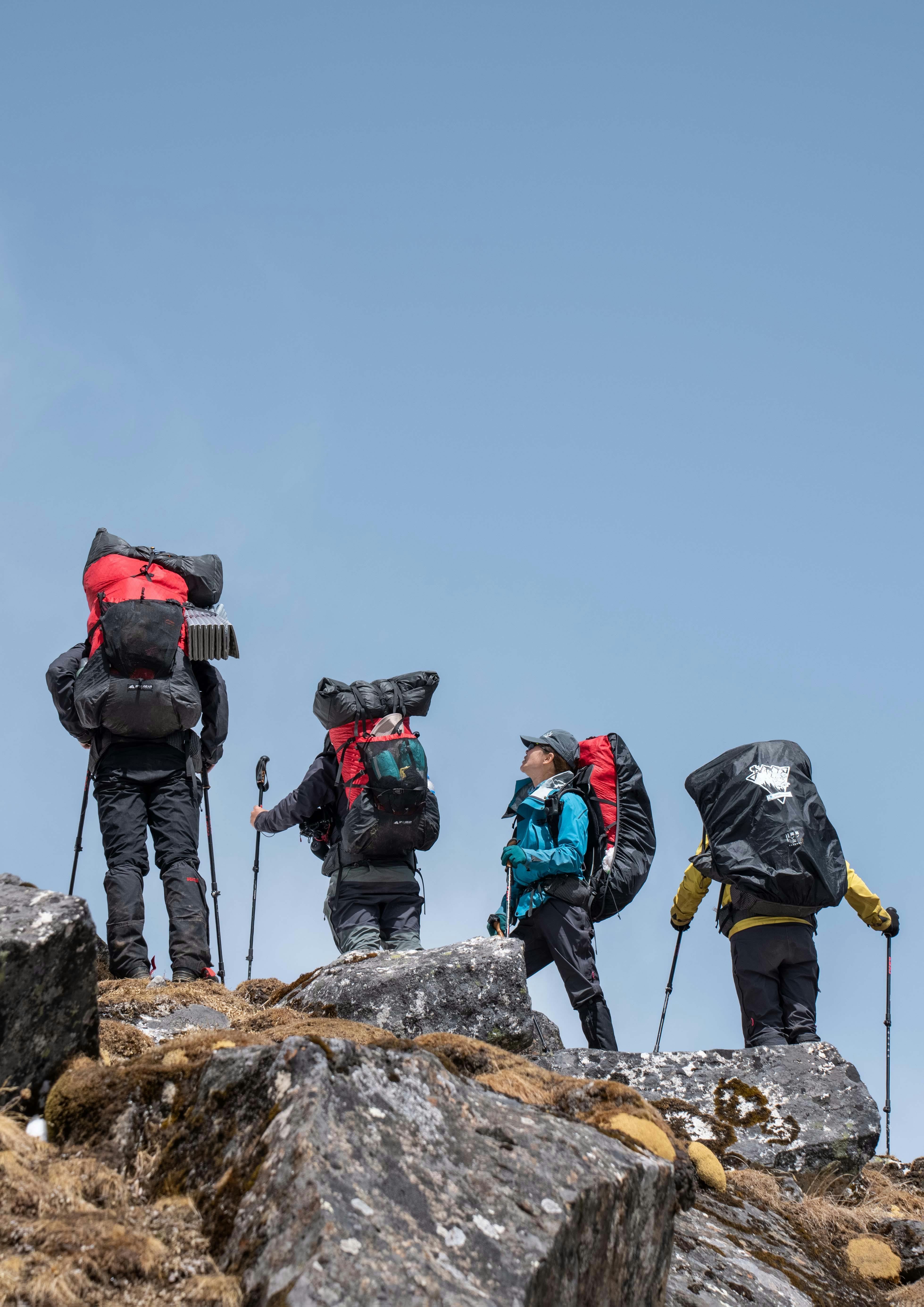 Four hikers with backpacks navigate a rocky mountain trail under a clear blue sky.