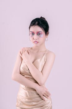 Portrait of a stylish woman standing gracefully in a satin dress showcasing vibrant makeup against a soft pastel background.