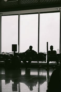 Silhouette of passengers waiting at an airport terminal with large glass windows and reflections.