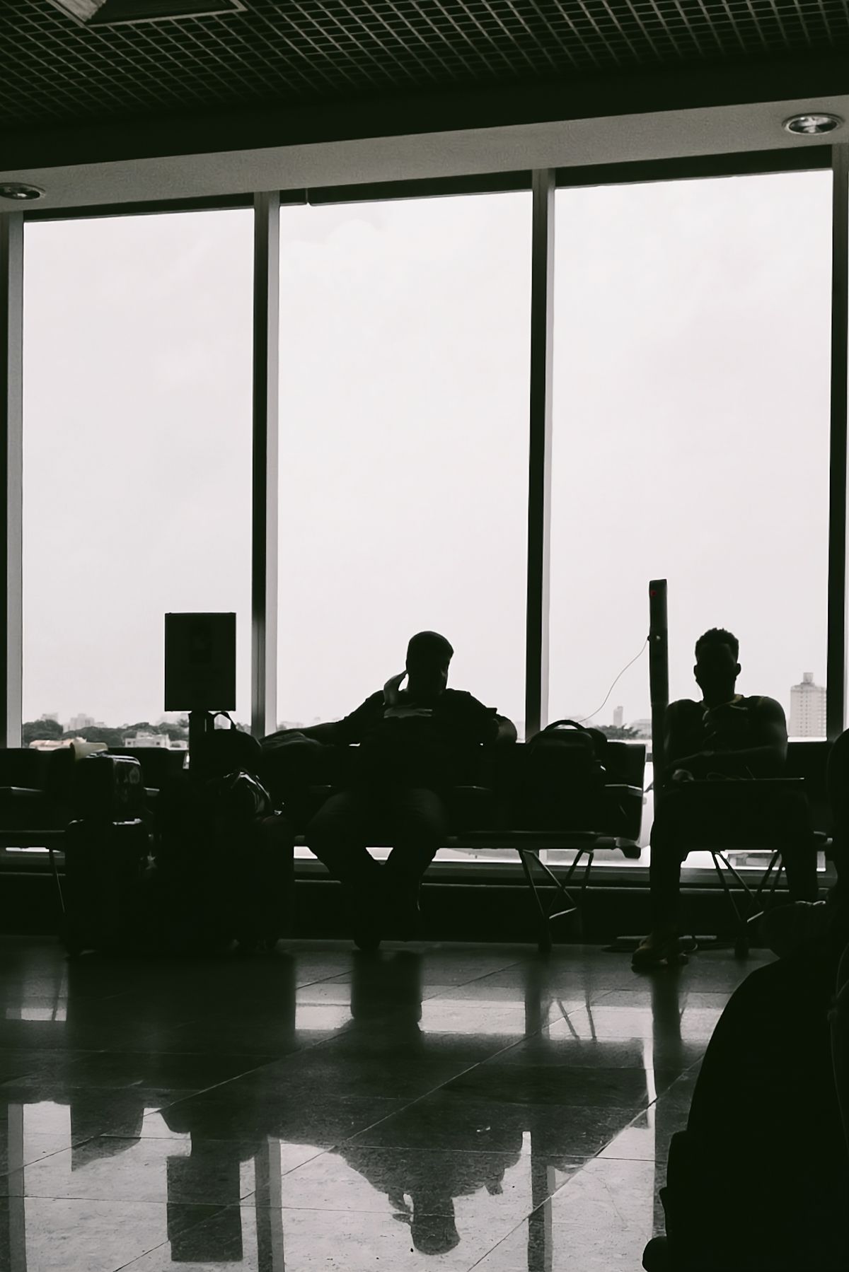 Silhouettes of two travellers sitting with their luggage by large terminal windows