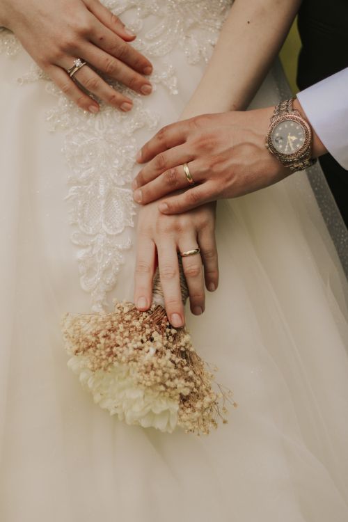 Free A close-up image of a married couple's hands displaying wedding rings and a bouquet on a wedding dress. Stock Photo