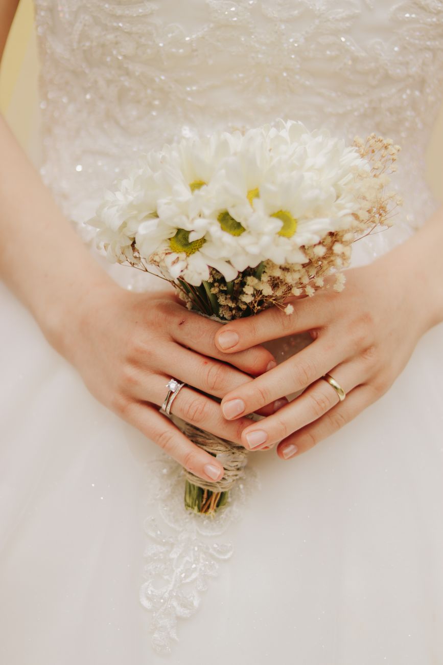 Close-up of a bride in a wedding dress holding a delicate white bouquet with rings visible.