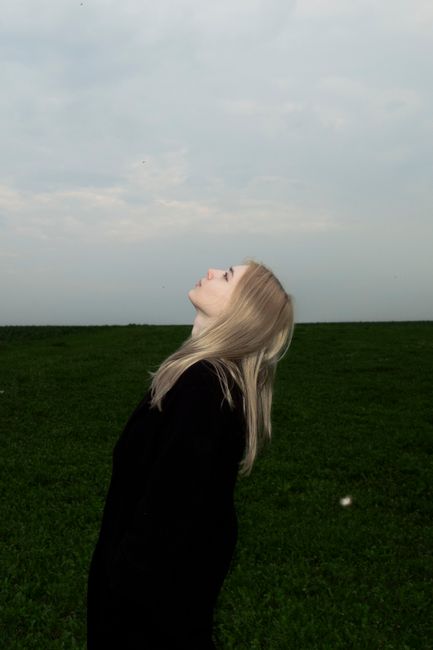 A young woman in a black coat looks upward in a serene outdoor setting, evoking a sense of contemplation and freedom.