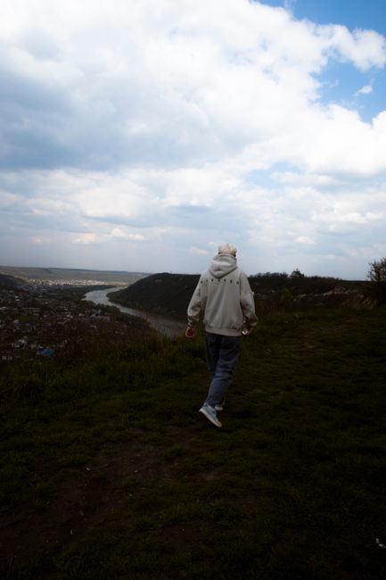 A young man in a grey hoodie walking along a hilly trail under a cloudy sky.