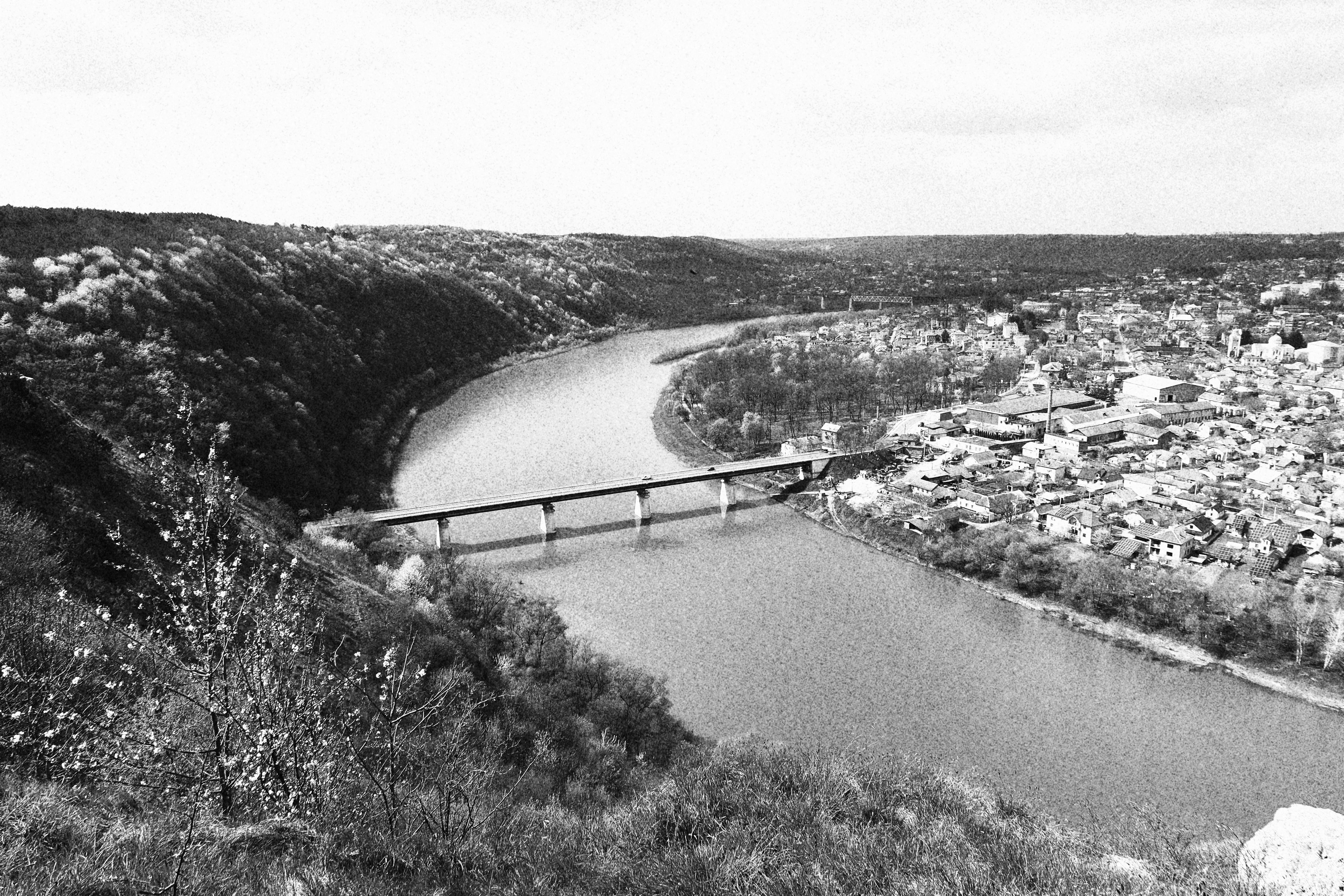 A black and white high angle view of a bridge spanning a river with a town and trees.
