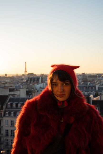 A woman in a red coat poses on a Paris rooftop with the Eiffel Tower in the background.