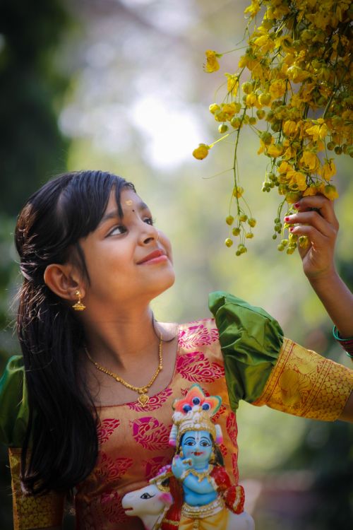 Free A young girl in traditional attire admires yellow flowers while holding a deity statue, symbolizing Indian culture. Stock Photo