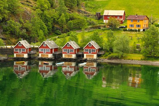 Charming red cabins reflecting on a serene lake, surrounded by lush greenery in a rural Nordic setting.