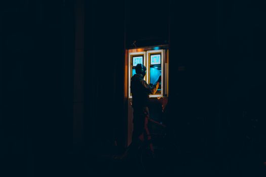 A man interacting with an ATM at night in a dark urban environment in Rotterdam.