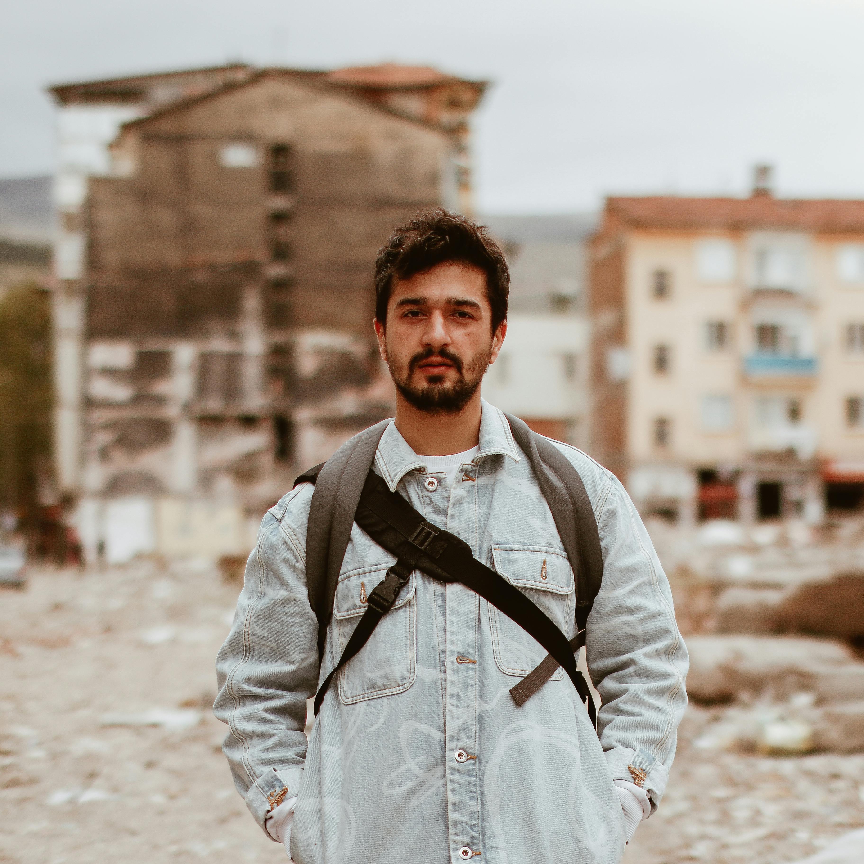 A man with a backpack in a denim jacket stands on a city street, showcasing urban exploration.