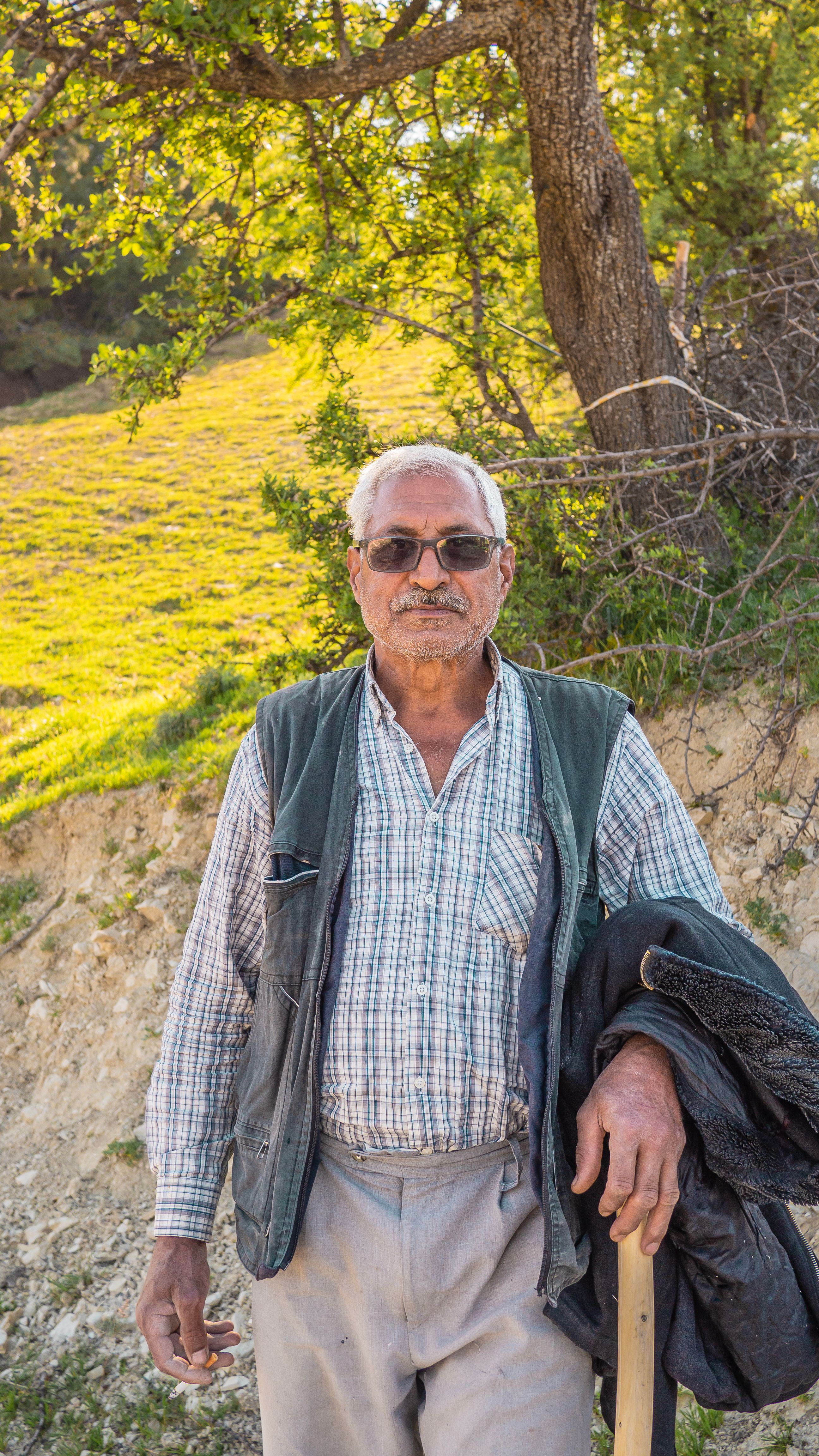 Portrait of an elderly man standing outdoors on a sunny day, in a rural setting.