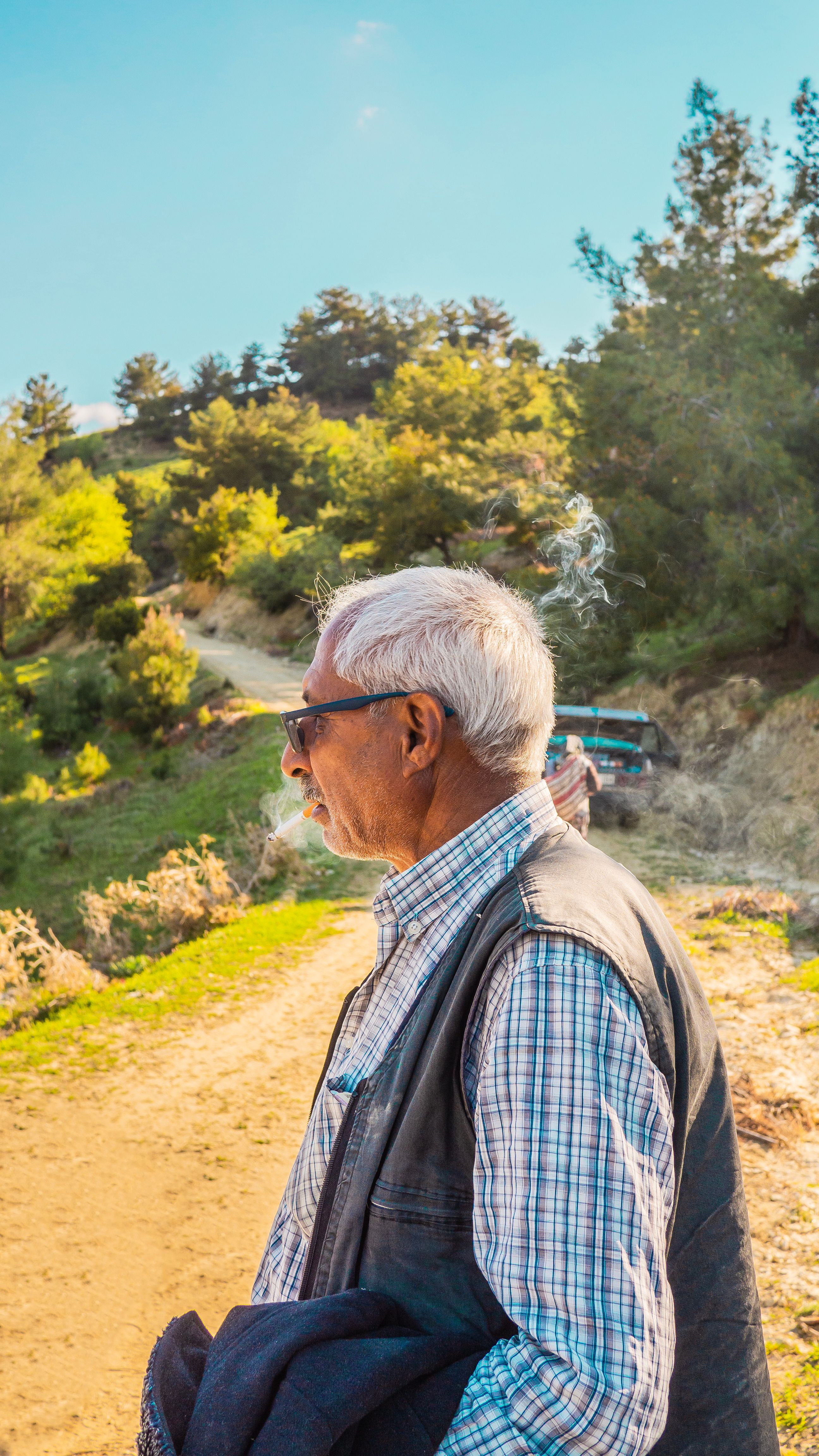 Senior man enjoying a sunny day, standing outdoors in a serene countryside area, surrounded by lush greenery.