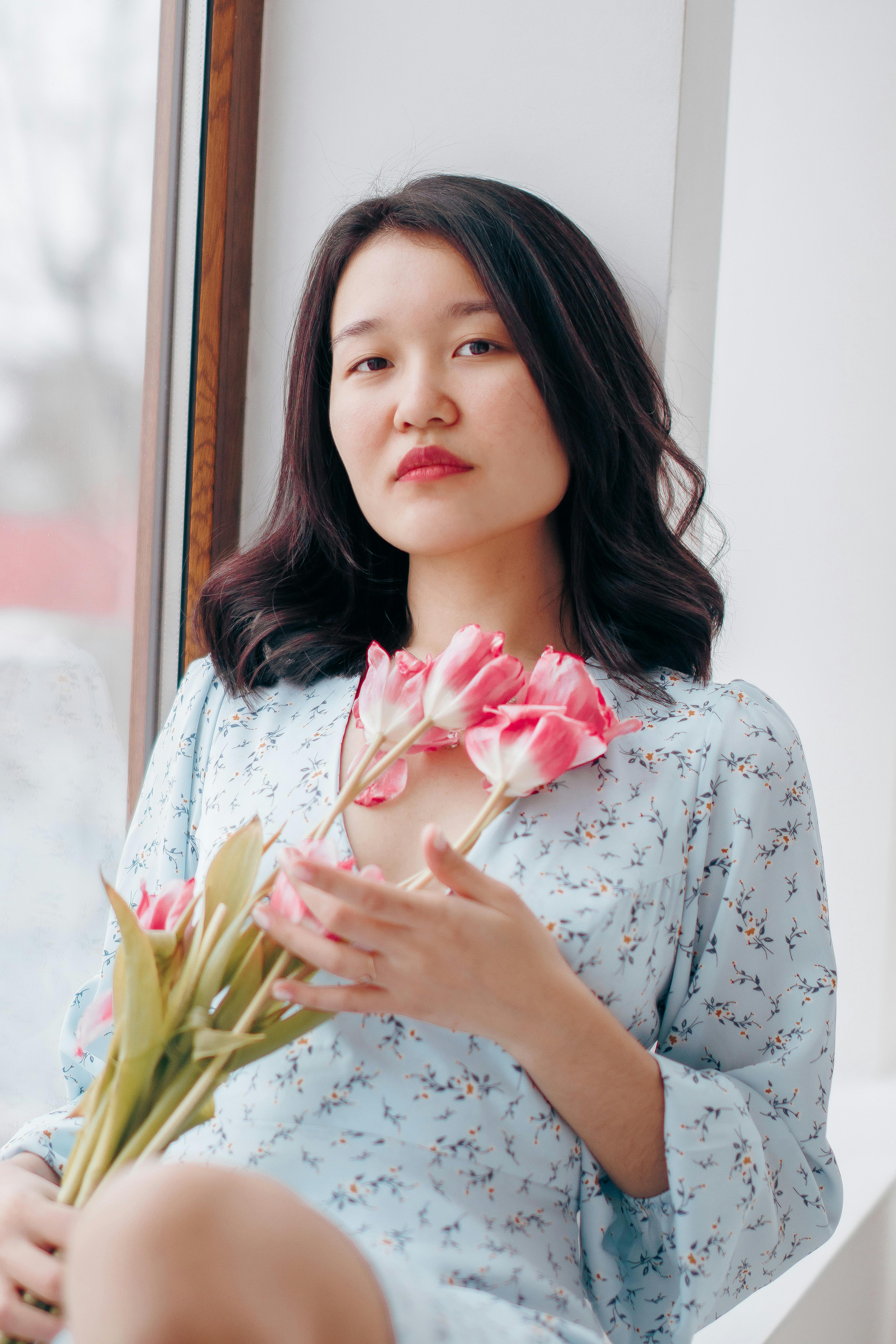 Asian woman in blue floral dress holding pink tulips by a window, exuding calm and elegance.