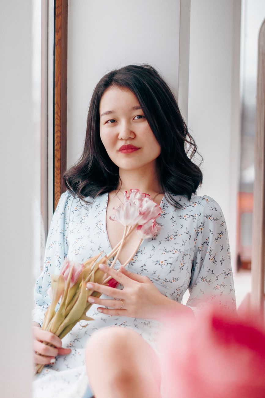Young woman in a floral dress holding tulips by a window, exuding elegance and tranquility.